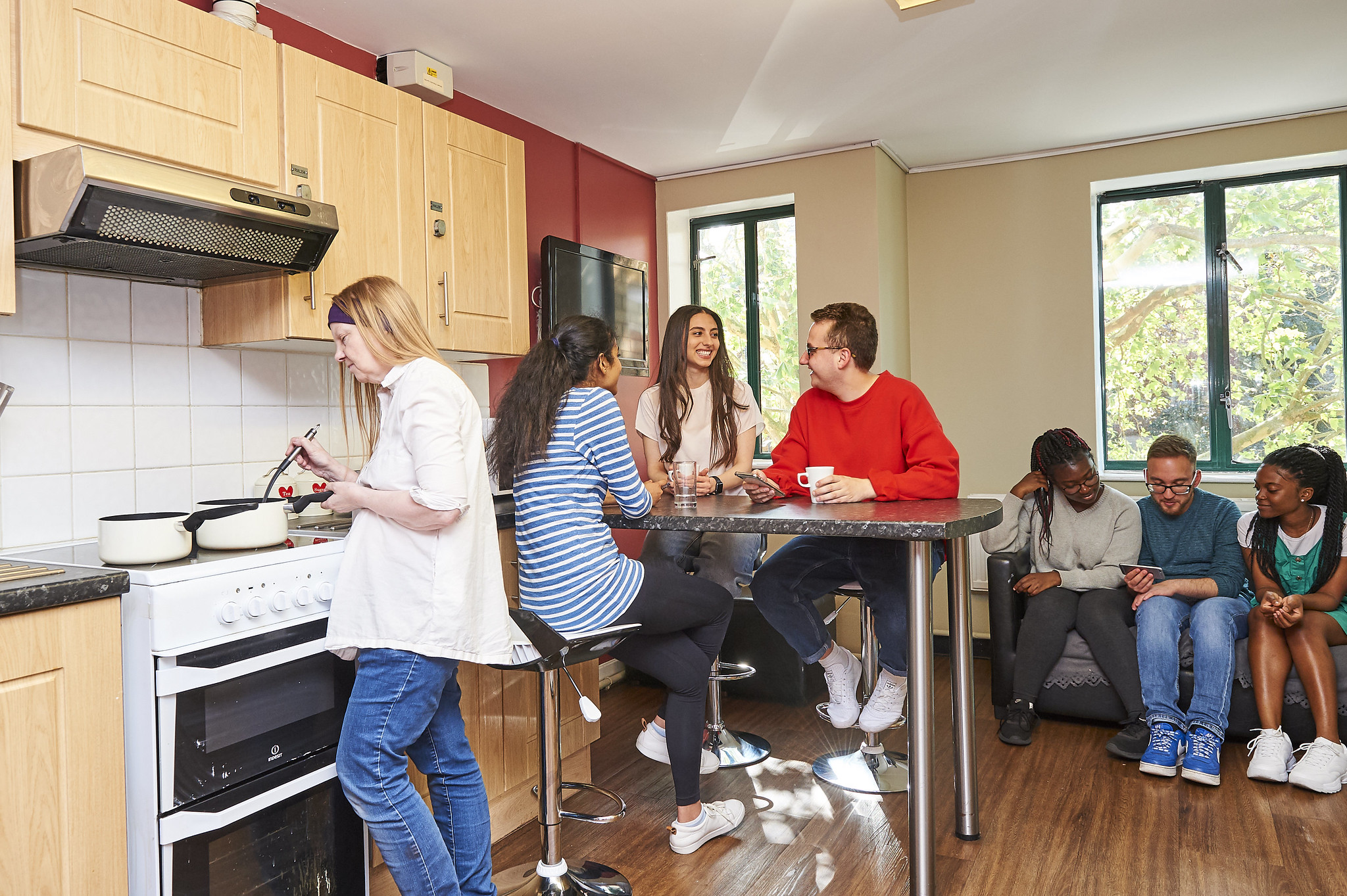 A group of students in a communal kitchen area, sitting around a table or on a sofa, one student working at the hob in the kitchen