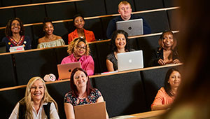 Students seated in a lecture hall, many with laptop computers, all facing the lecturer.